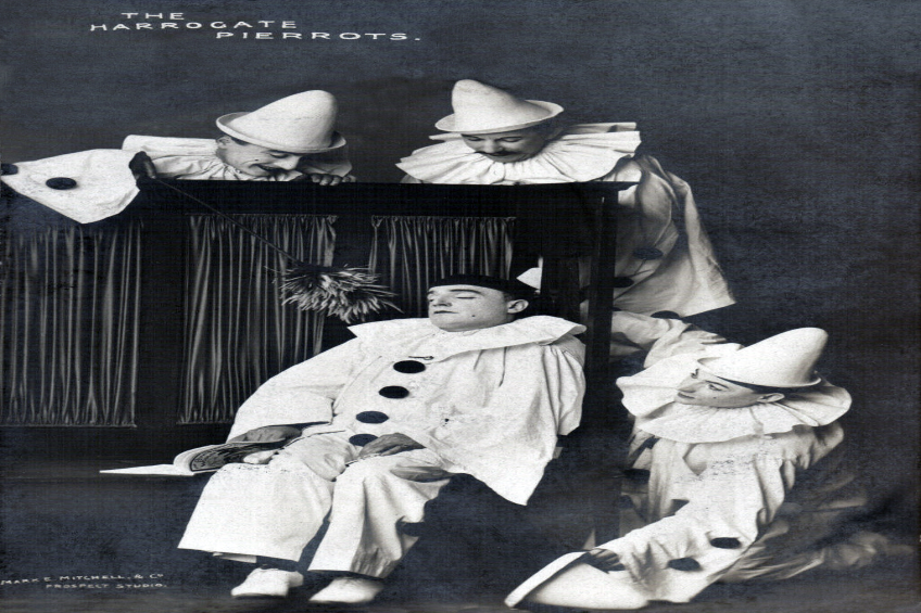 Ein Schwarz-Weiß-Foto einer Gruppe von Clowns, die auf einer Bühne sitzen, wobei eine Person ein Buch hält und Vorhänge im Hintergrund zu sehen sind.