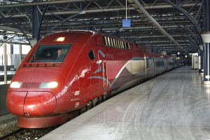 Ein roter Hochgeschwindigkeitszug steht auf einem Bahnhof mit einer Plattform auf der rechten Seite, einige Menschen stehen und Pfeiler und Stangen im Hintergrund.