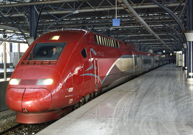 Ein roter Hochgeschwindigkeitszug steht auf einem Bahnhof mit einer Plattform auf der rechten Seite, einige Menschen stehen und Pfeiler und Stangen im Hintergrund.