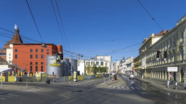 Stadtstraße mit Gebäuden, Straßeninfrastruktur, fahrenden Fahrzeugen, Fußgängern, Bäumen und Himmel.