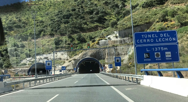 Auto fährt durch einen Tunnel auf einer Straße mit Geländern, Schildern, Polen, Verkehrszeichen, Bäumen, Felsen und einem klaren blauen Himmel.