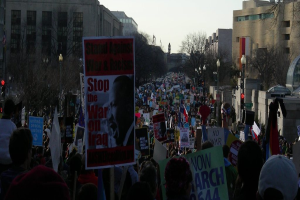 Menschen mit Plakaten auf einer Straße protestierend, mit Gebäuden, Bäumen, Laternen und dem Himmel im Hintergrund.