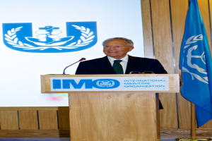 Eine Person steht hinter einem Podium mit einem Mikrofon, Brille und Text, der spricht, mit einer Wand im Hintergrund, auf der ein Logo und eine Flagge zu sehen sind.
