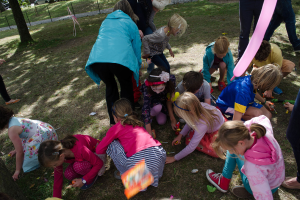 Eine Gruppe von Kindern in Hockstellung und einige stehende Menschen vor einem Ballon, mit einem Baumstamm, einem Zaun und einem geparkten Fahrzeug im Hintergrund.