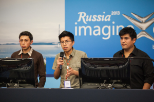 Drei Jungs stehen neben einem Schreibtisch mit zwei Computersystemen, einer spricht in ein Mikrofon, tr√§gt ID-Karten mit roten Markierungen, vor einer Wand mit einer Werbetafel, auf der "Russia Imagine 2013" steht.