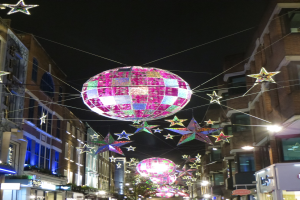 Eine festlich geschmückte Stadtstraße bei Nacht mit zahlreichen Lichtern und Dekorationen, darunter eine große Diskokugel an einem Gebäude.