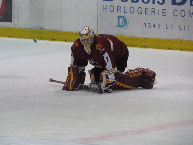 Eishockeyspieler in rot-gelber Uniform, der einen Schuss hält, trägt Helm, Handschuhe und Knieprotektoren und hält einen Eishockeyschläger, mit einer Wand und Text im Hintergrund.