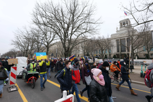 Eine große Gruppe von Menschen nimmt an einer Protestdemo auf einer Straße in Washington, D.C. teil, wobei einige Schilder und Transparente halten und andere Fahrräder fahren, sowie Schilder, Bäume und einen klaren blauen Himmel im Hintergrund.