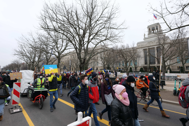 Eine große Gruppe von Menschen nimmt an einer Protestdemo auf einer Straße in Washington, D.C. teil, wobei einige Schilder und Transparente halten und andere Fahrräder fahren, sowie Schilder, Bäume und einen klaren blauen Himmel im Hintergrund.