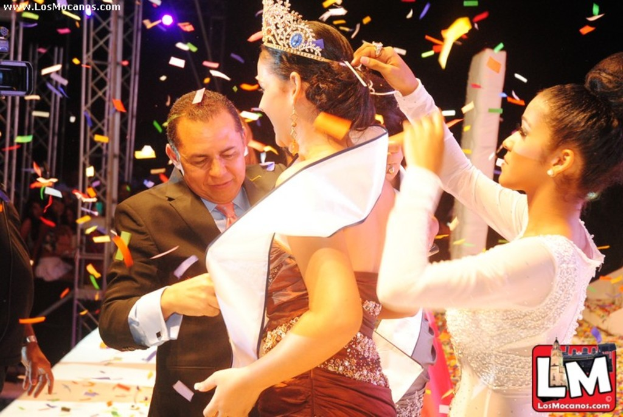 Ein Mädchen auf der Bühne bei einer Miss-World-Show mit einer Krone, mit einem anderen Mädchen, das sie hinter ihr anpasst, und einem Mann in einem schwarzen Anzug, der vor ihr die Hand schüttelt, vor einem dunklen Hintergrund mit Metallrahmen.