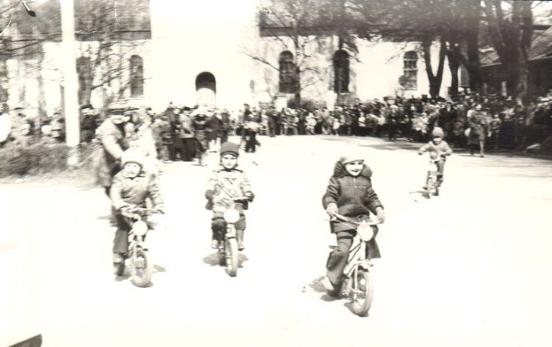 Schwarze-weiß-Foto von Kindern auf Fahrrädern, die eine Straße hinunterfahren, mit einem Gebäude und Bäumen im Hintergrund.