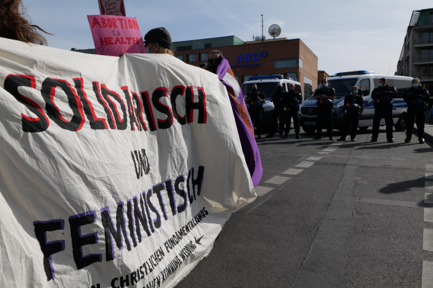 Gruppe von Menschen marschiert die Straße entlang und hält eine 'Solidarität und Feminismus'-Fahne, mit geparkten Fahrzeugen, Gebäuden, einer Schüsselantenne und einem klaren blauen Himmel im Hintergrund.