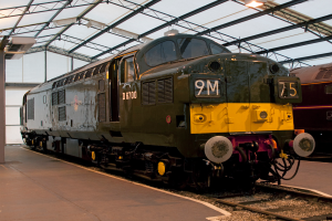 Ein Zug befindet sich auf den Schienen mit einem anderen Zug neben einer Plattform auf der rechten Seite; die linke Plattform hat einen Mast mit Metallstangen und Lampen.