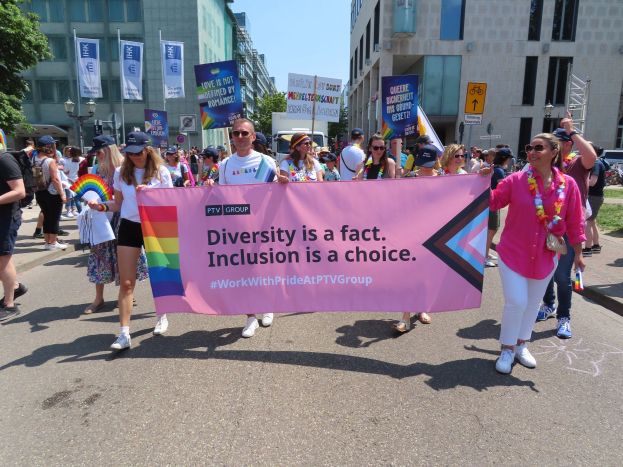 Eine bunte Gruppe von Menschen marschiert auf einer Straße, hält ein Banner mit der Aufschrift 'Diversität ist eine Tatsache, Inklusion ist eine Wahl', wobei einige eine Mütze und eine Schutzbrille tragen und andere zusätzliche Banner halten, vor einem Hintergrund von Gebäuden, Bäumen und einem klaren blauen Himmel.