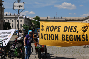 Eine Gruppe von Menschen steht vor einem Gebäude und hält ein gelbes Banner hoch, auf dem "Hope Dies, Action Begins" steht, mit Fahrrädern in der Nähe und einem Pfahl mit einer Tafel daran, unter einem klaren blauen Himmel mit Bäumen im Hintergrund.