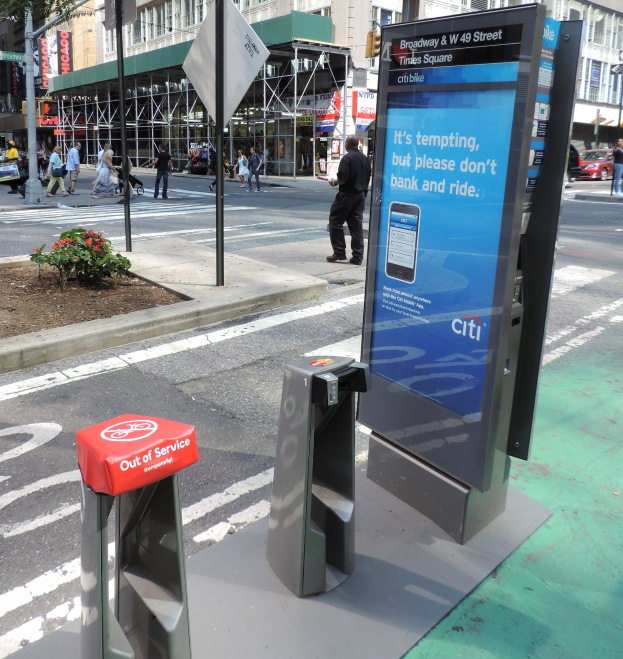 Eine belebte Stadtstraße mit Fußgängern auf dem Gehweg, Fahrzeugen auf der Straße und einer Bushaltestelle mit dem Schild "Citi" und einem Mülleimer in der Nähe, vor Häusern, Bäumen und Ampeln.