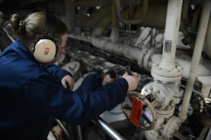 Zwei Frauen in blauen Uniformen arbeiten an einer Maschine in einer Öl- und Gasindustrieumgebung, wobei eine ein Objekt hält und Kopfhörer trägt, und Rohre im Hintergrund sichtbar sind.