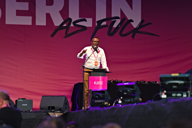 Ein Mann in einem weißen Hemd und braunen Hosen steht auf einer Bühne und hält ein Mikrofon in der Hand, hinter ihm eine pinkfarbene Fahne mit der Aufschrift 'Berlin as Fuck' und eine Menge vor ihm.