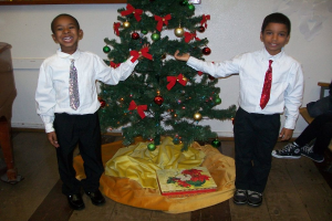 Ein geschmückter Weihnachtsbaum steht in einem Raum mit einer Kiste und anderen Gegenständen auf dem Boden, einem hölzernen Tisch links, einem Stuhl rechts und zwei Jungen in weißen Hemden, die lächeln und den Baum halten.