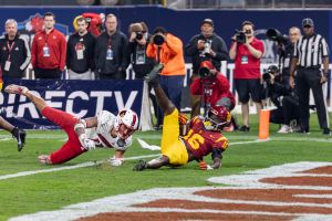 Ein Fußballspieler taucht in die Endzone ein, um einen Touchdown zu erzielen, mit einer Gruppe von Menschen am Boden, einige halten Kameras, und ein Banner mit Text im Vordergrund, während Geländer, Pfosten und andere Gegenstände im Hintergrund sichtbar sind.