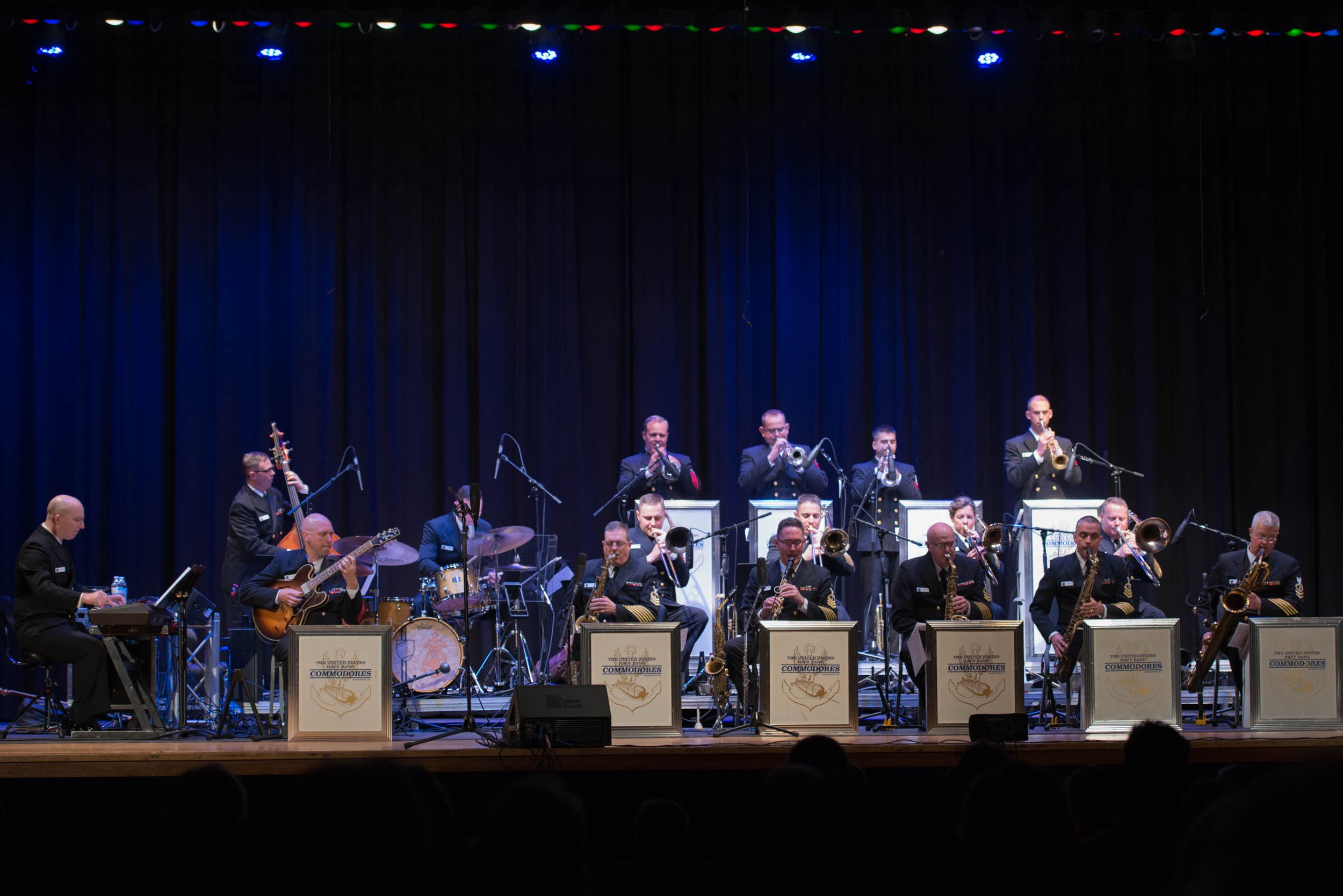Eine Gruppe Musiker spielt auf einer hell erleuchteten Bühne, während "The New Orleans Jazz Band" auf Schildern hinter ihnen zu sehen ist und das Publikum darunter zusieht.