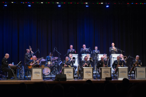 Eine Gruppe Musiker spielt auf einer hell erleuchteten Bühne, während "The New Orleans Jazz Band" auf Schildern hinter ihnen zu sehen ist und das Publikum darunter zusieht.