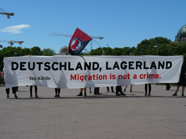 Eine Gruppe von Menschen mit einem Banner, auf dem "Deutschland, Lagerland Migration ist kein Verbrechen" steht, und einer Flagge, die auf dem Boden steht mit Straßenlaternen, Bäumen, Kränen, einem Gebäude und einem bewölkten Himmel im Hintergrund.