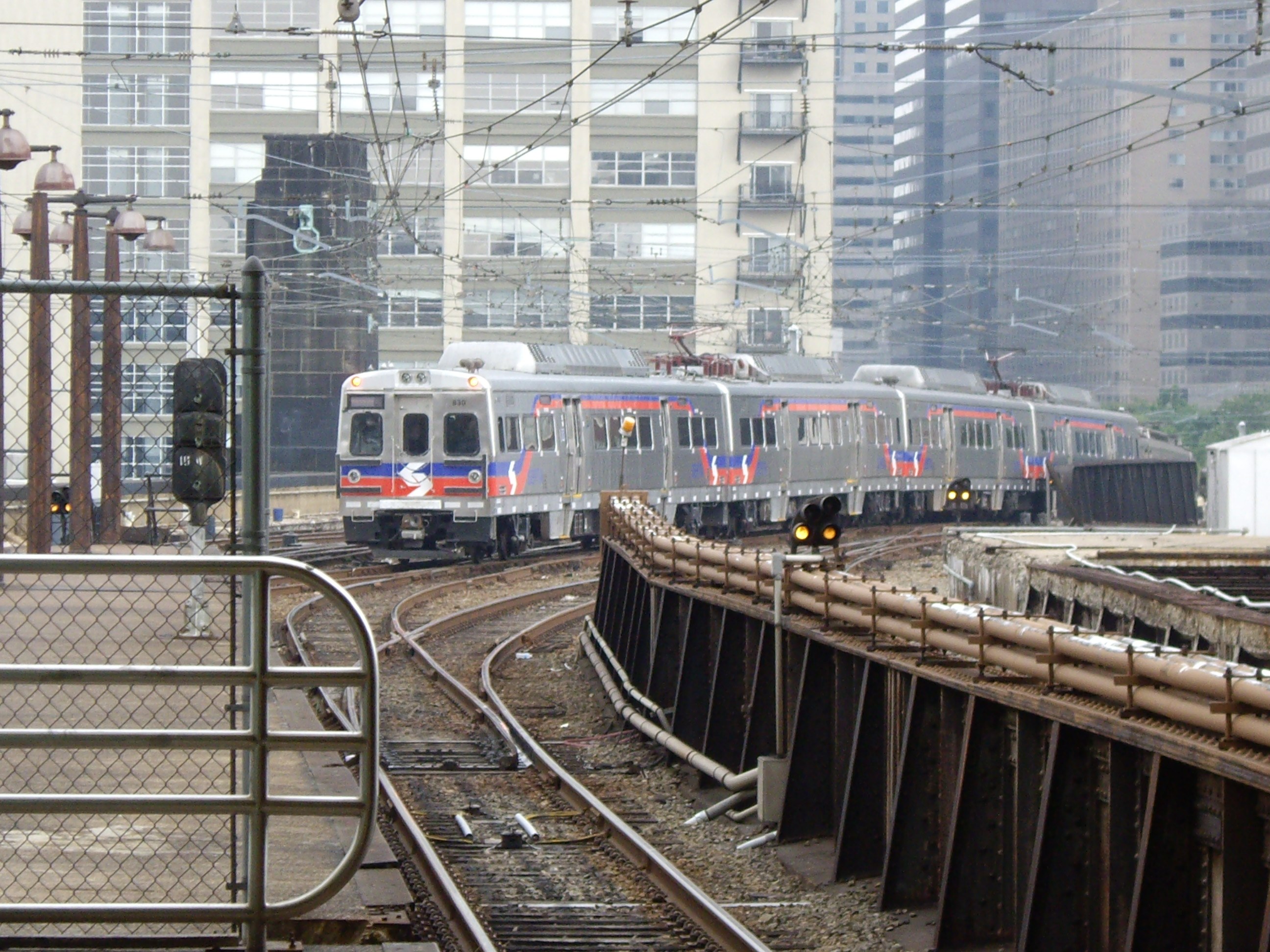 Nahverkehrszug fährt neben Hochhäusern mit sichtbarer Bahninfrastruktur einschließlich Gleise, Masten, Lampen und Verkehrsschildern.
