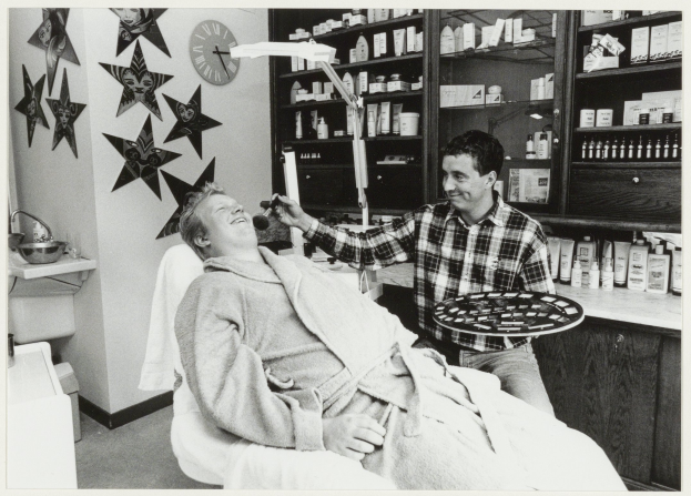 Ein Mann lächelt in einem Zahnarztstuhl, während ein anderer Mann daneben steht und ein Tablett hält, mit einem Waschbecken, Sternen, einer Uhr und Regalen mit Flaschen im Hintergrund.