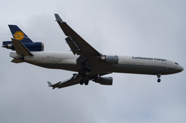 Ein Lufthansa Cargo Boeing 777-300ER im Flug, mit ausgebreiteten Tragflächen und aktivierten Triebwerken, mit sichtbarem Text auf der Rühlung gegen einen bewölkten Himmel.