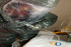 Eine Person in einem Laborkittel, Helm und Handschuhen injiziert eine Substanz in eine Flasche auf einem Tisch mit Papieren und einer Waage.