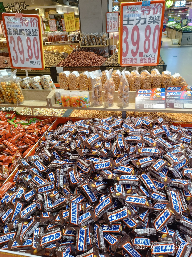 Vitrine mit verschiedenen Süßigkeiten, einschließlich Snickers, und Lebensmitteln in einem Geschäft mit Deckenbeleuchtung und Hintergrundobjekten.