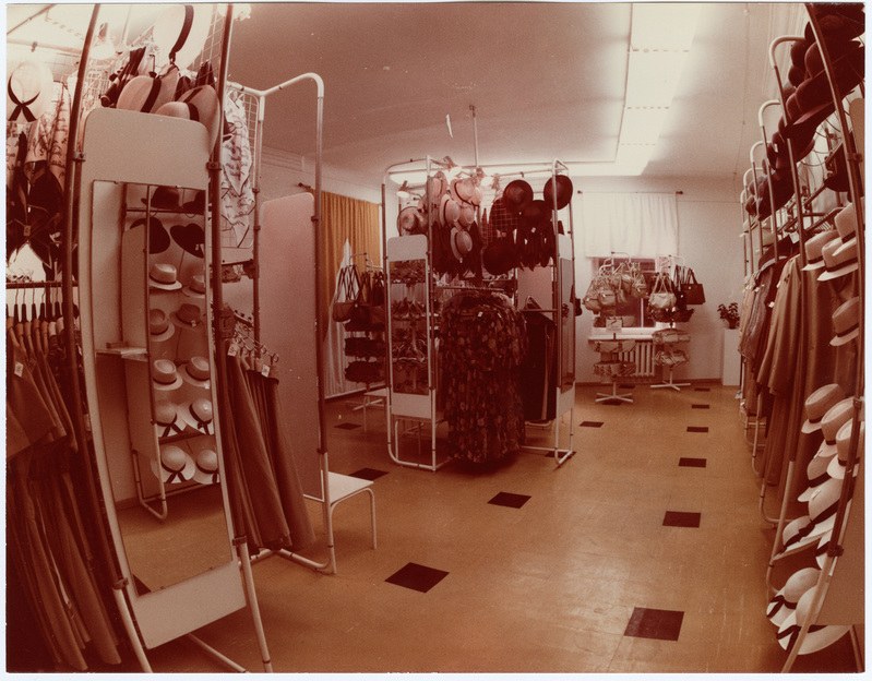 Altes Foto eines Bekleidungsgeschäfts-Innenraums mit Kleidung an Bügeln, Hüten auf Ständern, einem Spiegel, einem Stuhl, einem Tisch mit Gegenständen, Vorhängen und Deckenleuchten.