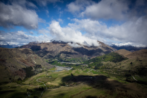 Ein Panoramablick von einem Berggipfel in Queenstown, Neuseeland, der saftiges grünes Gras, Bäume, eine gewundene Straße und einen Himmel voller weißer, flauschiger Wolken zeigt.