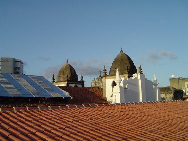 Eine Stadtansicht mit mehreren Gebäuden im Vordergrund unter einem blauen Himmel, mit Solarpanelen auf einem Dach, die den Einsatz erneuerbarer Energien anzeigen.