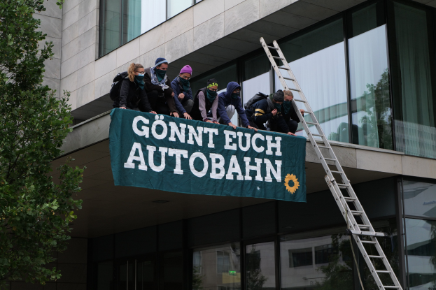 Gruppe von Menschen auf einem Gebäudedach mit einem Banner, mit einem Baum links und Glasfenstern im Hintergrund, die zu protestieren scheinen.