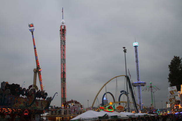 Eine Menschenmenge versammelt sich um einen hell erleuchteten Faschingsumzug in der Nacht, mit beleuchteten Fahrgeschäften, Pfählen und Bäumen, unter einem bewölkten Himmel.