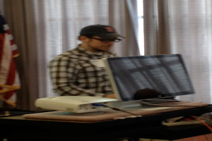 Ein Mann mit Mütze und Brille steht vor einem Computersystem, im Hintergrund ist eine Flagge zu sehen.