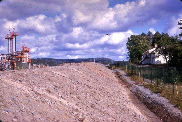 Ein großer Schmutzhaufen auf einem umzäunten Gebiet mit Gras, Pflanzen, Bäumen und einem Haus, mit einer Ölraffinerie links und Hügeln und einem bewölkten Himmel im Hintergrund.