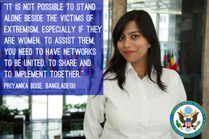 Eine Frau in einem weißen Hemd steht vor einem Gebäude mit einer Inschrift über Einheit und Unterstützung für Opfer von Extremismus, insbesondere Frauen. Ein Logo ist in der rechten unteren Ecke sichtbar.