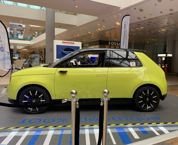 Ein gelbes wasserstoffbetriebenes Elektroauto vor einem Gebäude mit Absperrpollern, Bannern, einer Person im Auto, einer Gruppe von Menschen, Schildern, einer Topfpflanze, Deckenleuchten und Lüftern.