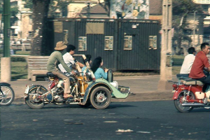 Mehrere Motorradfahrer auf einer Straße mit einem Gehweg, einem Pfahl, einer Bank und einem Baumstamm dahinter und Gebäuden, einer Hütte und Gras im Hintergrund.