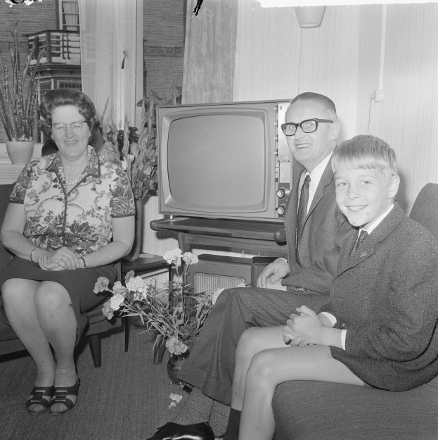 Schwarzes und weißes Foto einer Familie aus drei Personen - einem Mann in einem Blazer und Krawatte, einer Frau und einem Jungen - die auf einem Sofa sitzen, mit Blumenvasen auf einem nahen Tisch, einem Fernseher vor ihnen und Vorhängen im Hintergrund.