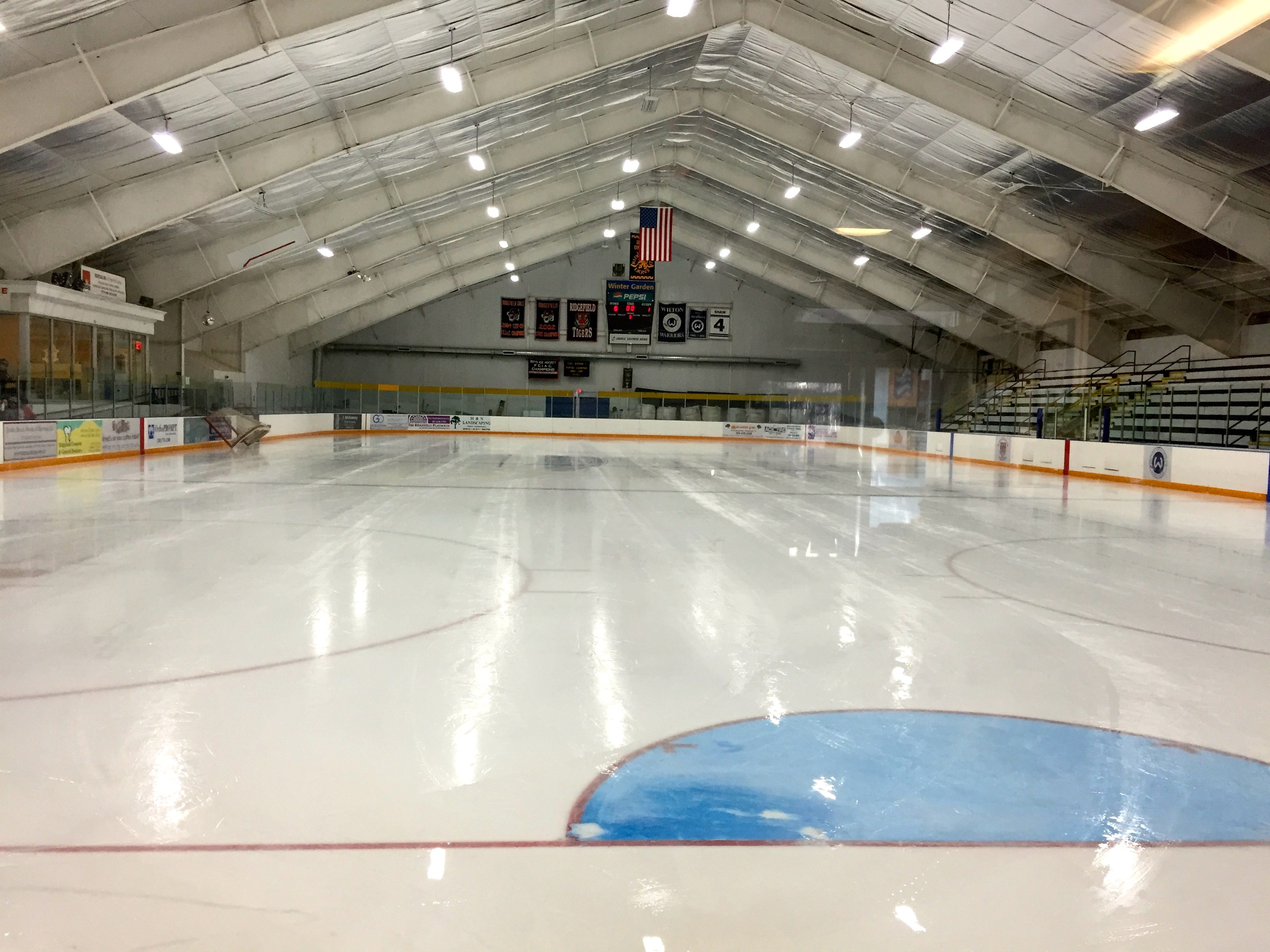 Große Indoor-Eisfläche mit Deckenbeleuchtung, Umrandungsplanken, einer Rückwand, Treppen mit Geländern und einigen Menschen, wahrscheinlich eine Hockeyanlage.