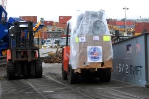 Ein Mann bedient einen Gabelstapler, der eine große Kiste trägt, umgeben von verschiedenen Fahrzeugen, Menschen, Metallobjekten, Containern, Polen und Bäumen unter einem klaren blauen Himmel.