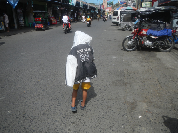 Ein junger Junge geht eine Straße entlang, die von Motorrädern und anderen Fahrzeugen wimmelt und von Gebäuden mit Schildern gesäumt ist, unter einem klaren blauen Himmel mit Bäumen und Pfosten im Hintergrund.