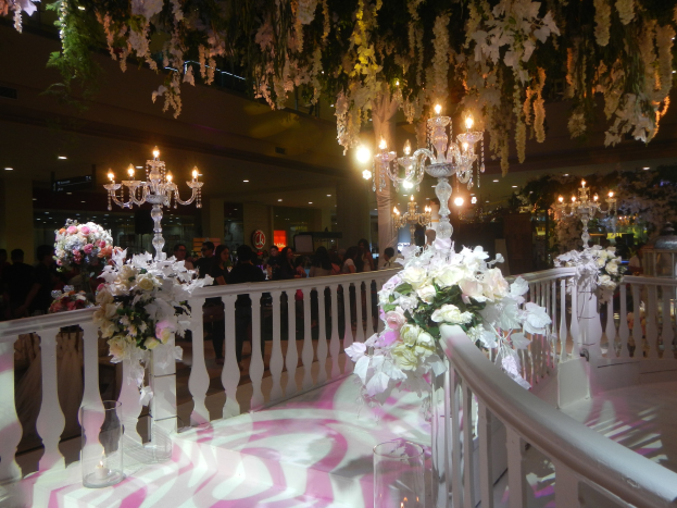 Eine Hochzeitszeremonie in einem großen Raum mit weißen und pinken Blumen und Kronleuchtern, mit Geländern, Blumenbouquets und Gläsern auf dem Boden, mit Menschen im Hintergrund.