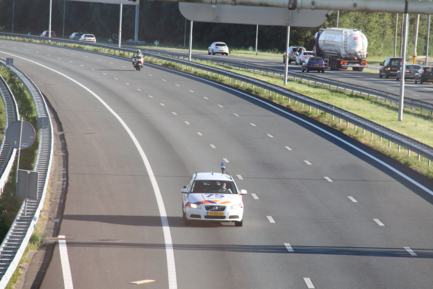Ein Auto auf einer Autobahn mit einem stationären Polizeiwagen am Rande, andere Fahrzeuge im Verkehr und Bäume und Straßeneinrichtungen im Hintergrund.