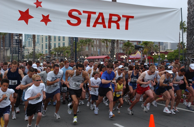 Gruppe von Menschen bei einem Marathon mit einem Verkehrskegel im Vordergrund und einer Fahne im Hintergrund, umgeben von Bäumen, Laternenmasten, Gebäuden und einem klaren blauen Himmel.