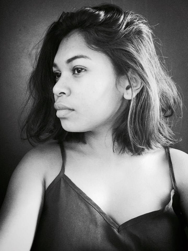 Eine selbstbewusste Frau in einem schwarzen Kleid posiert in einem Schwarz-Weiß-Foto, steht mit einer selbstbewussten Haltung vor einer Wand, ihr glatter Bob und dezentes Make-up unterstreichen ihren entschlossenen Ausdruck.